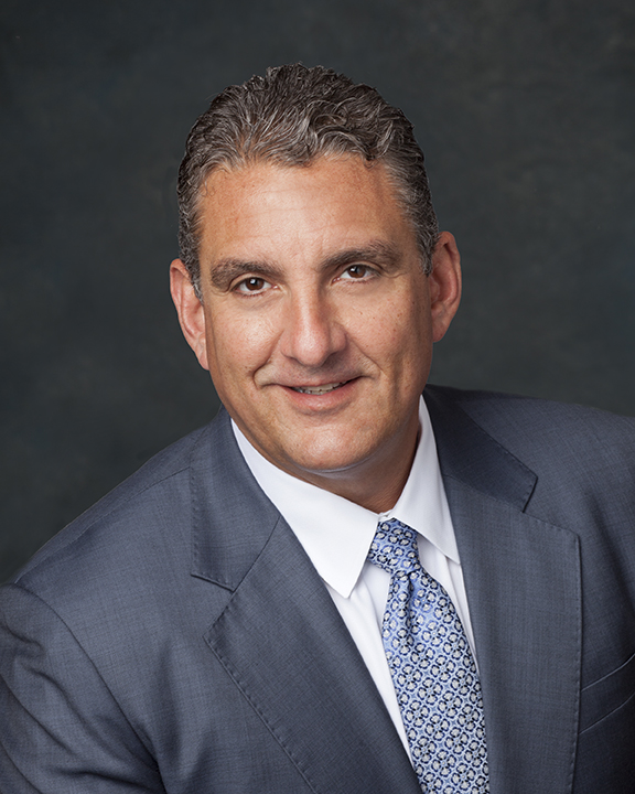 A man in a gray suit, white shirt, and patterned blue tie smiles at the camera against a dark, textured background.