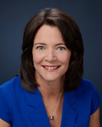 A woman with shoulder-length brown hair, wearing a royal blue blazer and a silver necklace, smiles at the camera against a plain dark blue background.