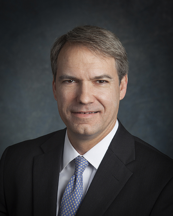 A middle-aged man with short gray hair, wearing a dark suit, white shirt, and patterned tie, is posing in front of a dark, textured background. He is looking at the camera and smiling slightly.