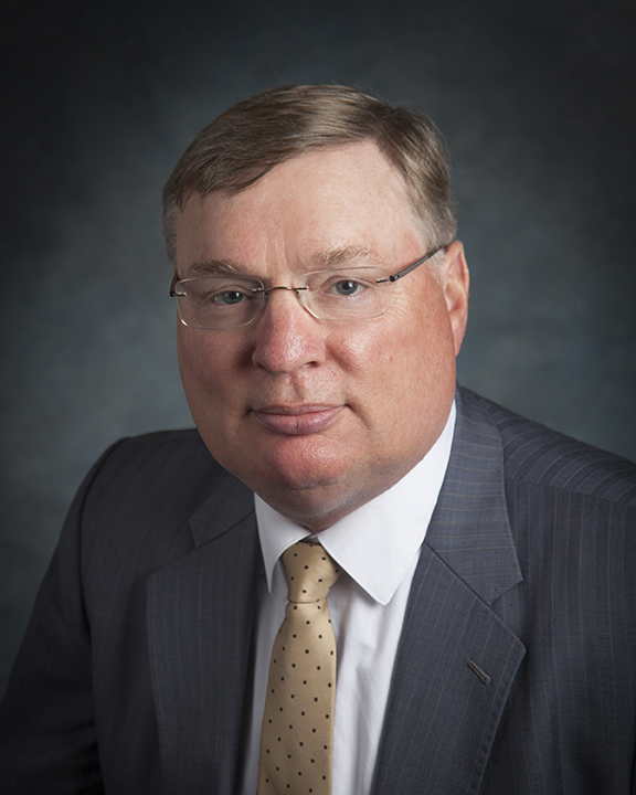 A middle-aged man with light skin, short light brown hair, and glasses, wearing a gray pinstripe suit, white shirt, and beige polka dot tie, posing against a dark, plain background.