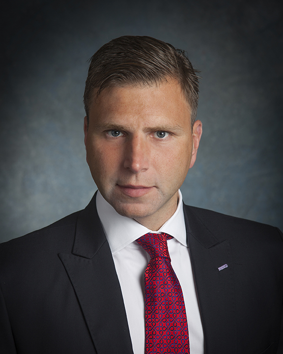 A man in a dark suit, white shirt, and red patterned tie poses for a professional headshot against a blue-gray textured background. He has short brown hair and a neutral expression.