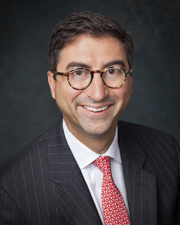 A man wearing glasses, a dark pinstripe suit, a white shirt, and a red patterned tie, smiling at the camera against a dark background.