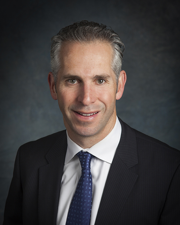 A man with short gray hair, wearing a dark pinstripe suit, white dress shirt, and blue patterned tie, poses for a professional headshot against a dark, textured background.
