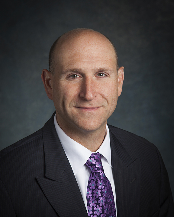 A man in a dark pinstripe suit, white shirt, and purple patterned tie is smiling slightly at the camera against a dark, textured background.