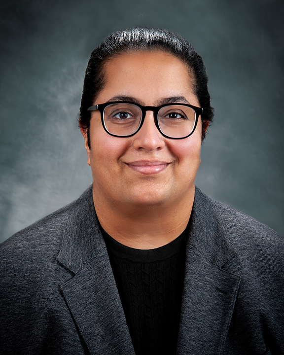 A person with dark hair pulled back, wearing black glasses, a dark sweater, and a gray blazer, smiles slightly in front of a gray textured background.