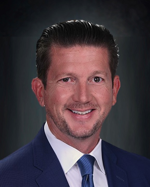 A man with short, slicked-back dark hair and trimmed facial hair, wearing a blue suit, white shirt, and blue tie, smiles in front of a dark, plain background.