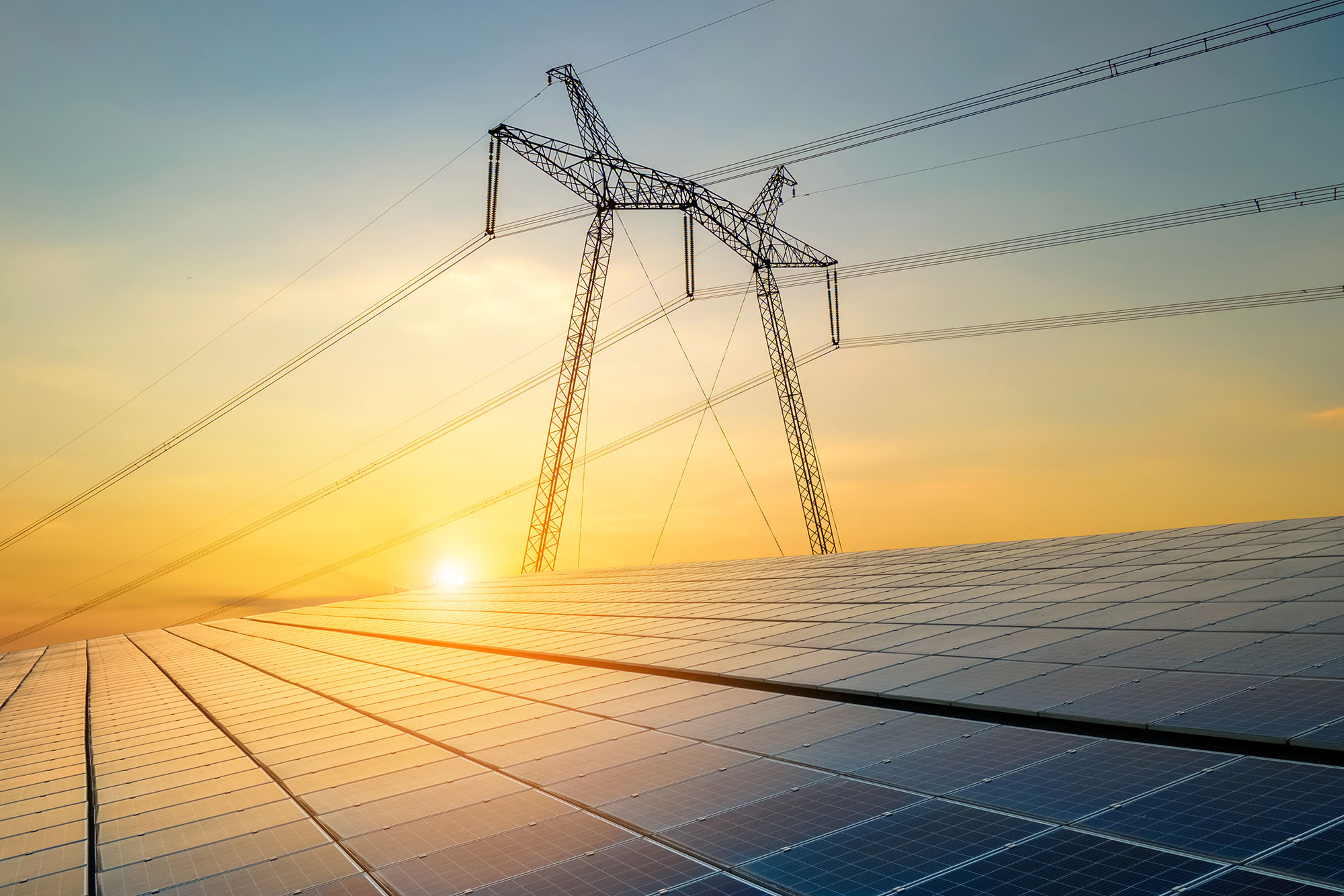 Rows of solar panels are shown in the foreground with a tall electricity transmission tower and power lines above, set against a bright sunset sky.