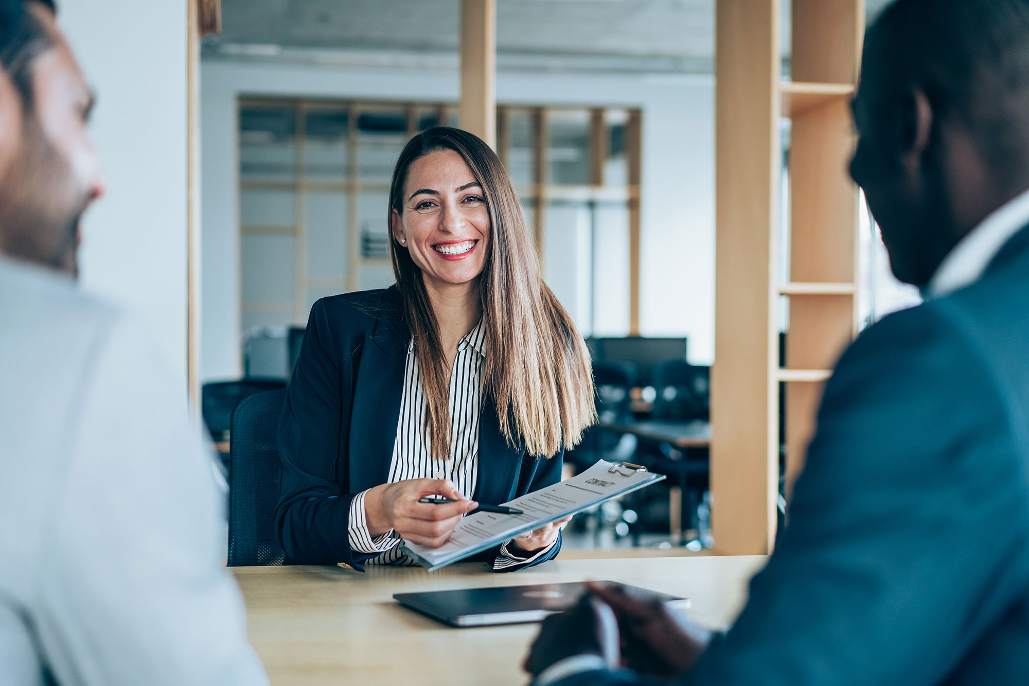 A woman in business attire smiles while holding a resume during a job interview, sitting at a table with two men in an office setting.