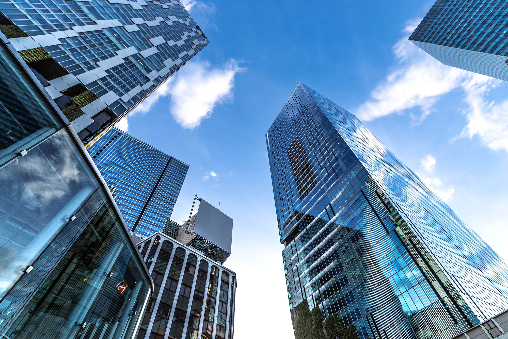Tall modern skyscrapers with glass facades reflect the blue sky and clouds, seen from a low angle. The buildings feature geometric patterns and sharp lines, emphasizing an urban cityscape.