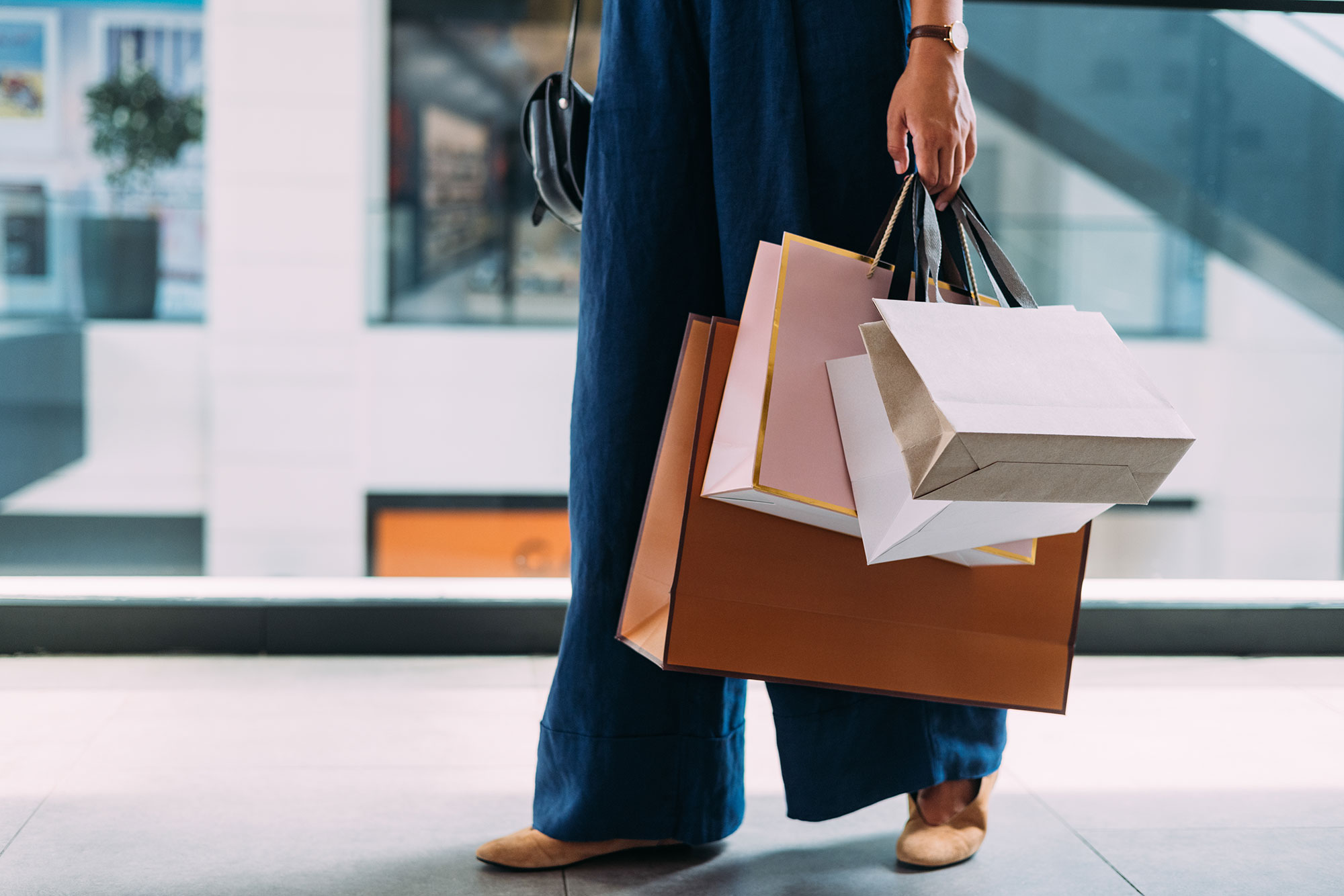 A person wearing blue pants and beige shoes is holding several shopping bags of different sizes and colors while standing indoors, possibly in a mall. Only the lower half of the body is visible.