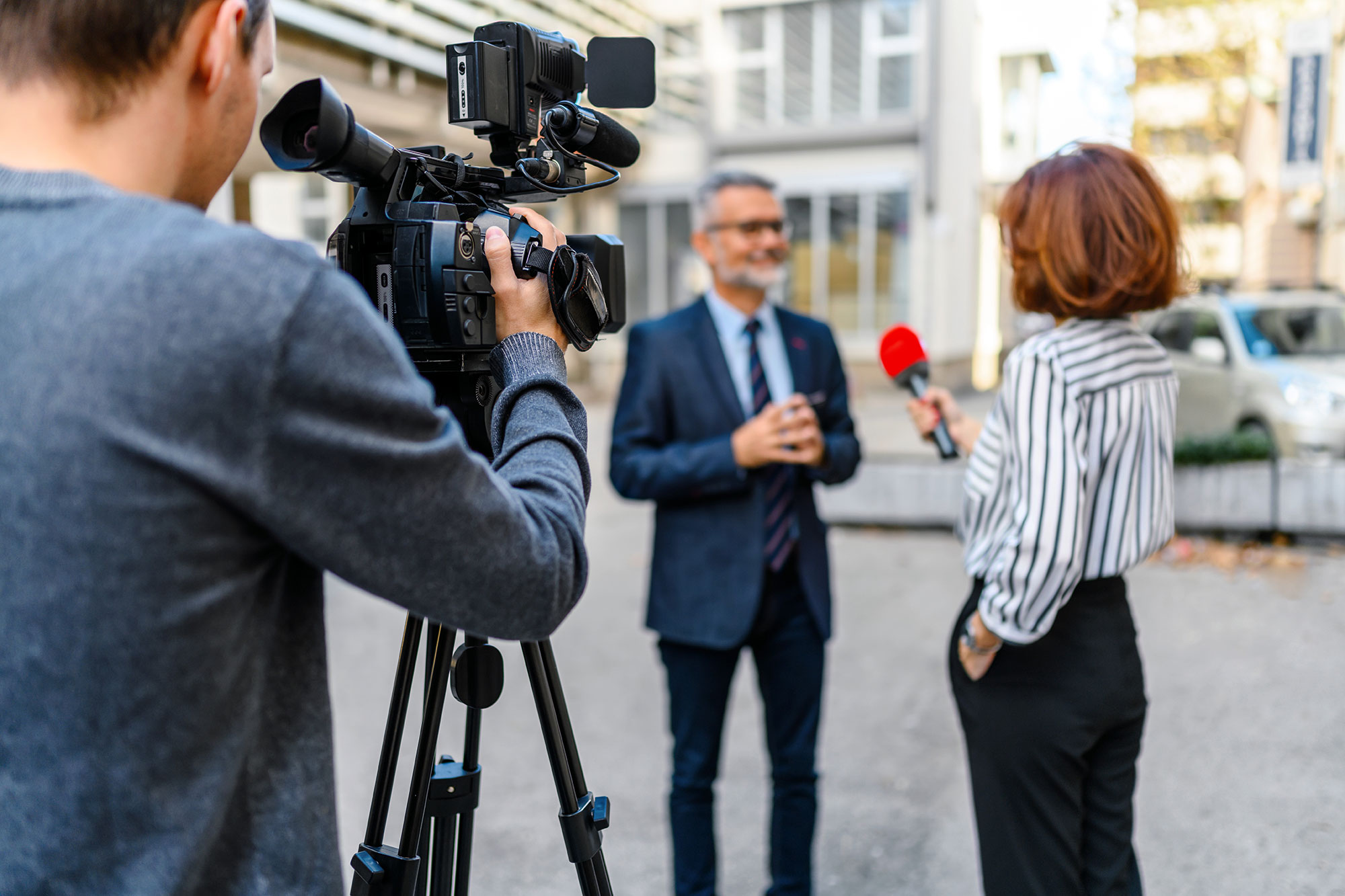 A camera operator films a man in a suit being interviewed by a woman holding a red microphone, outdoors in an urban setting with buildings and parked cars in the background.