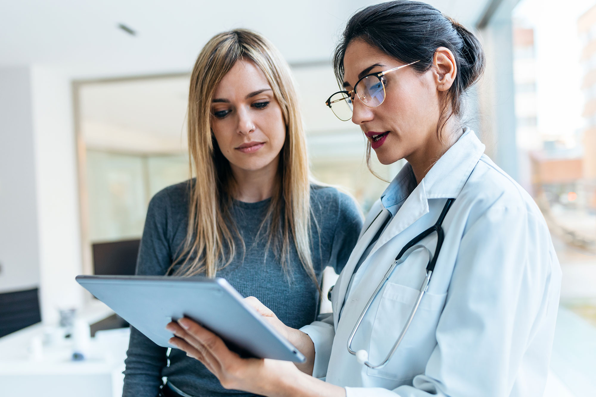 A doctor with a stethoscope shows a tablet to a patient. They are standing together in a bright, modern medical office, discussing information displayed on the tablet.