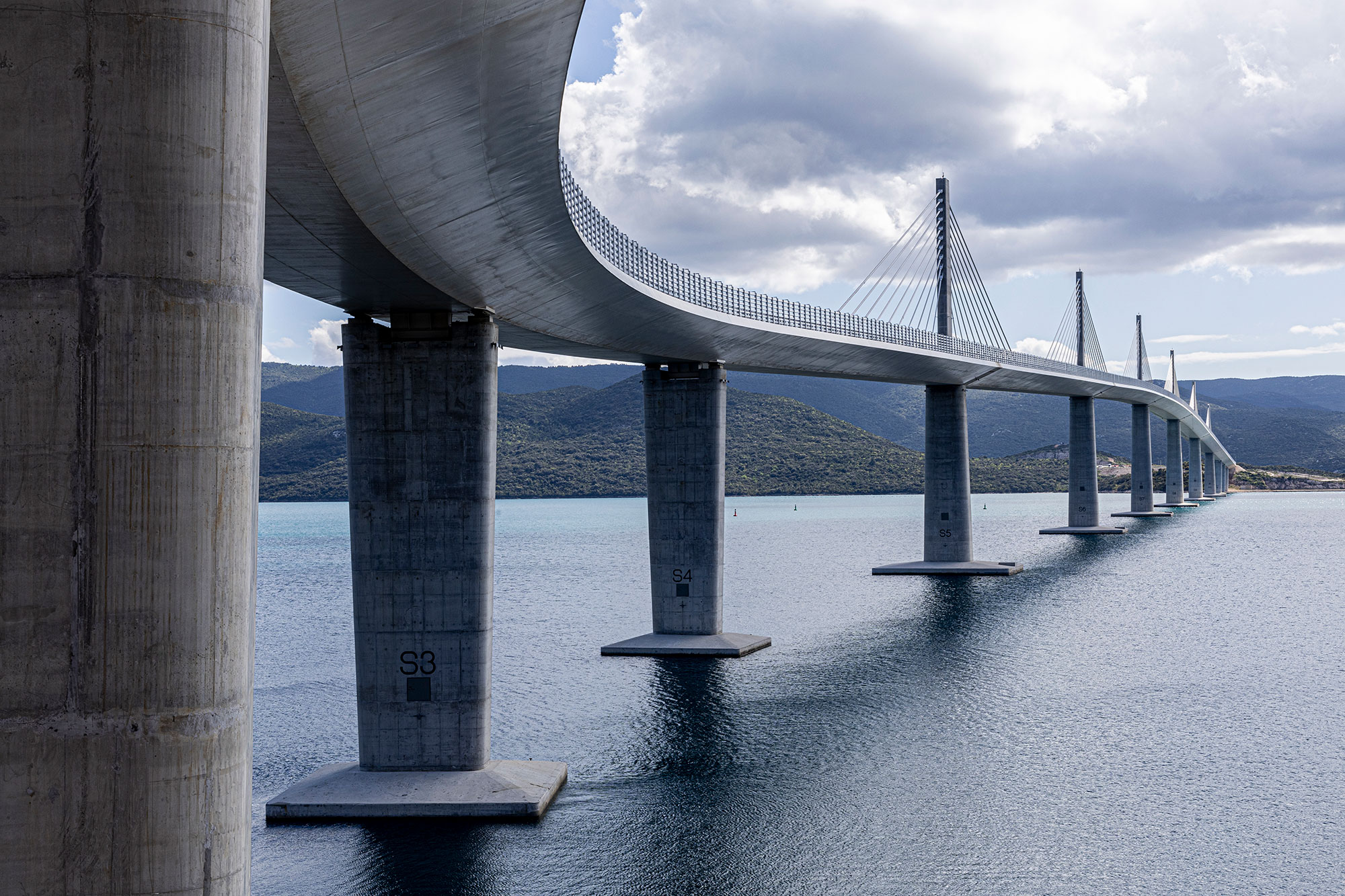 A modern cable-stayed bridge with tall concrete pillars spans across calm blue water, leading toward distant hills under a partly cloudy sky.
